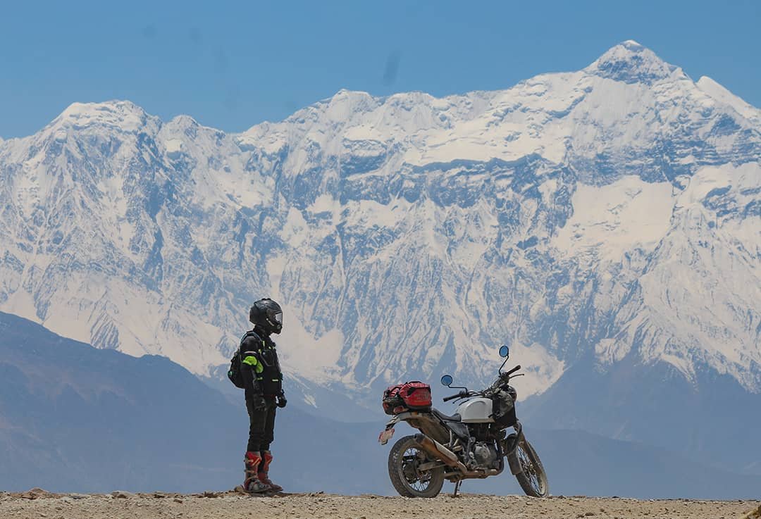 A rider stands beside his motorcycle facing a wall of snow-covered peaks